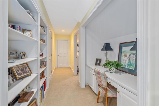 corridor featuring light colored carpet, a desk, and baseboards