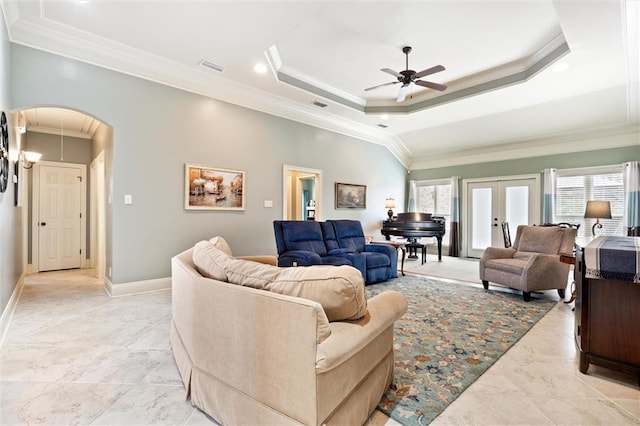 living room featuring baseboards, ceiling fan, a tray ceiling, ornamental molding, and arched walkways