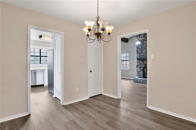 unfurnished dining area with ceiling fan with notable chandelier, dark wood-type flooring, a stone fireplace, and plenty of natural light