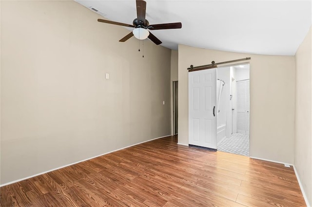 unfurnished bedroom featuring ensuite bathroom, a barn door, ceiling fan, lofted ceiling, and hardwood / wood-style flooring