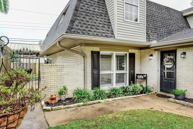 entrance to property featuring a shingled roof, mansard roof, fence, and brick siding