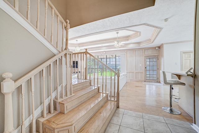 stairs with tile patterned floors, ceiling fan with notable chandelier, ornamental molding, and a tray ceiling