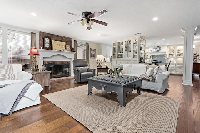 living room with a brick fireplace, dark wood-type flooring, ceiling fan, and ornate columns