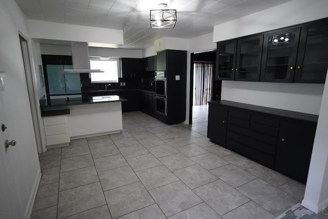 kitchen featuring a peninsula, a sink, light tile patterned flooring, island range hood, and dark cabinets
