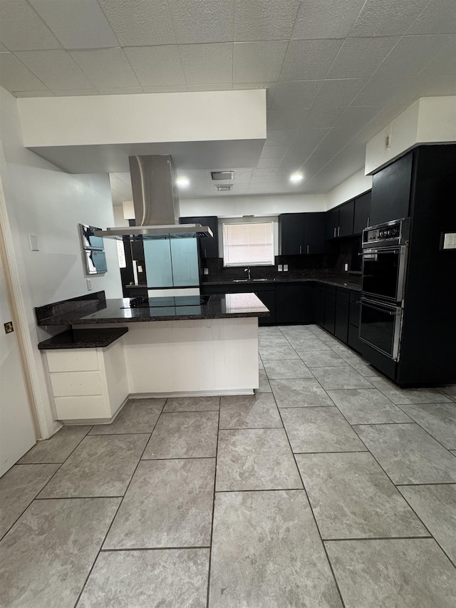 kitchen featuring double oven, dark stone counters, island range hood, sink, and kitchen peninsula