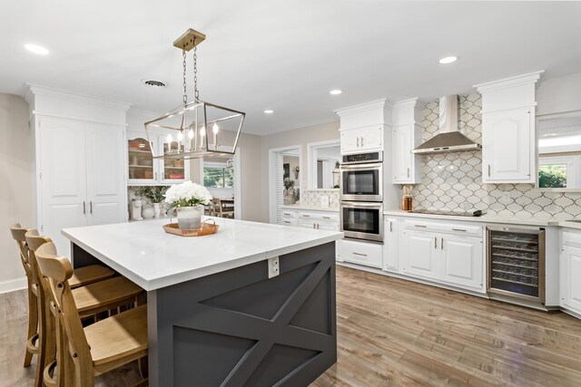 kitchen featuring tasteful backsplash, double oven, beverage cooler, wall chimney range hood, and white cabinets