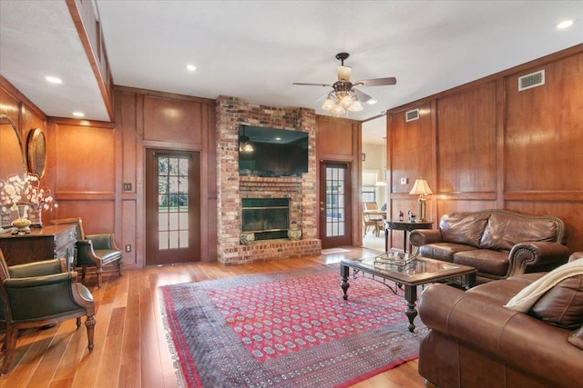 living room featuring a brick fireplace, wooden walls, ceiling fan, and light wood-type flooring