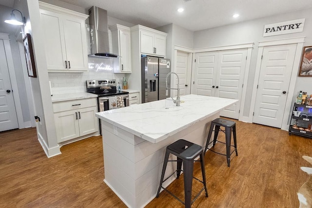 kitchen with wall chimney exhaust hood, white cabinetry, stainless steel appliances, and a kitchen island with sink