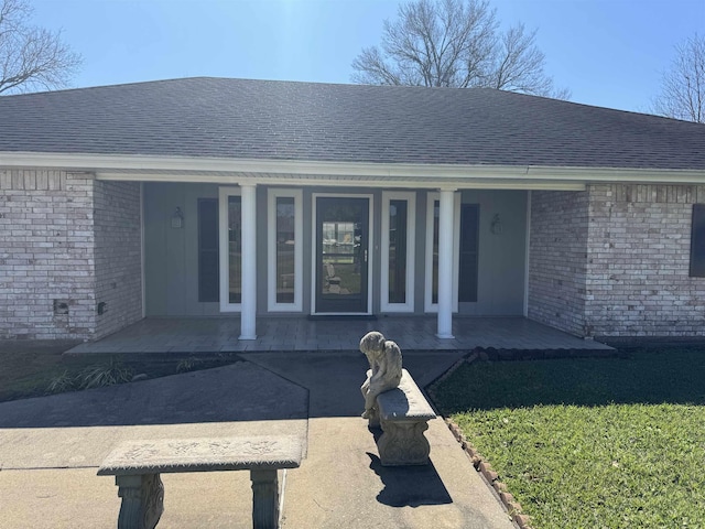 view of exterior entry with covered porch, brick siding, and roof with shingles