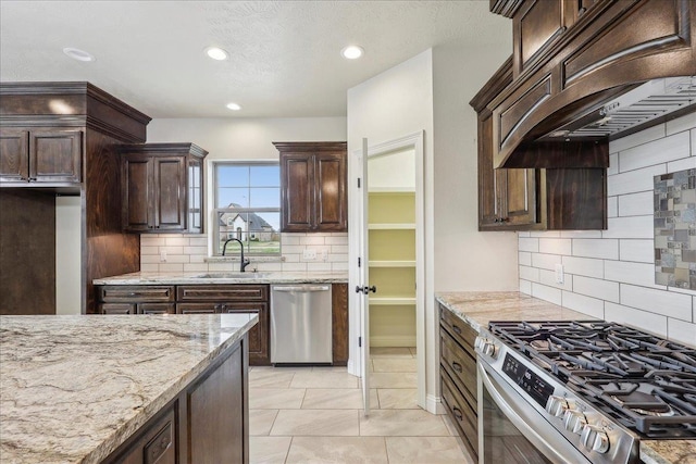 kitchen featuring sink, custom exhaust hood, dark brown cabinetry, stainless steel appliances, and light stone countertops