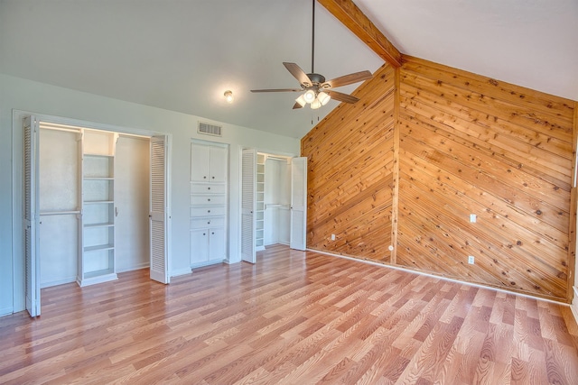 unfurnished bedroom with wooden walls, ceiling fan, multiple closets, light wood-type flooring, and vaulted ceiling with beams