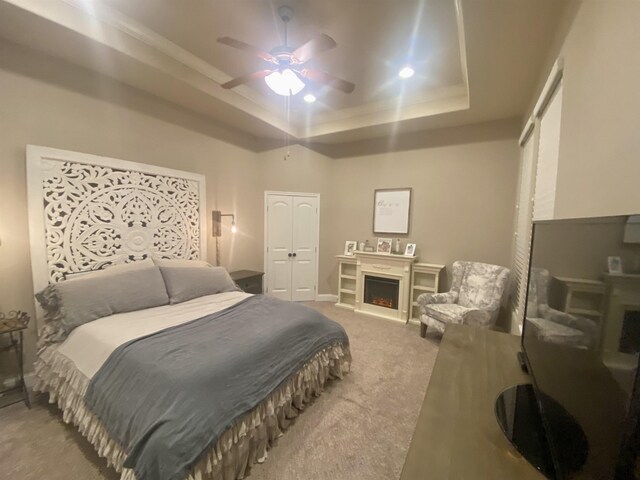 bedroom featuring a closet, a raised ceiling, carpet flooring, ceiling fan, and a warm lit fireplace