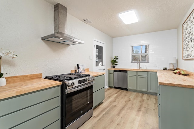 kitchen featuring wall chimney range hood, appliances with stainless steel finishes, a sink, green cabinets, and wood counters