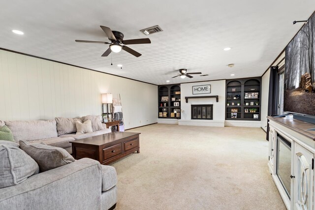 living area featuring visible vents, built in features, a glass covered fireplace, light colored carpet, and ceiling fan