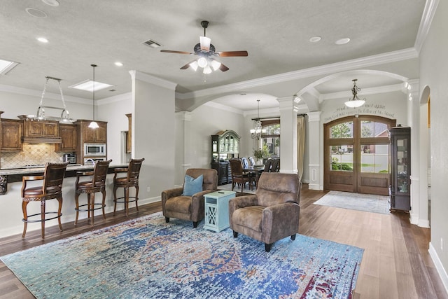 living room featuring wood-type flooring, ornamental molding, a textured ceiling, ceiling fan with notable chandelier, and ornate columns