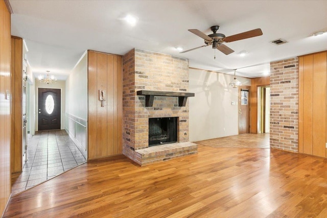 unfurnished living room with wood walls, a fireplace, ceiling fan with notable chandelier, and light wood-type flooring