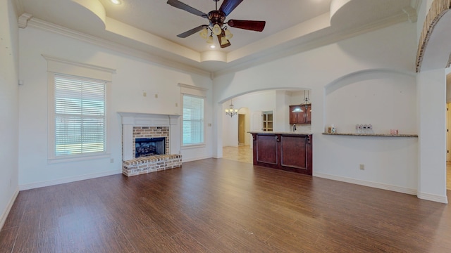 unfurnished living room with a brick fireplace, a raised ceiling, wood finished floors, and ceiling fan with notable chandelier