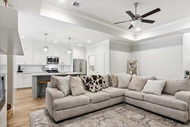 living room featuring a raised ceiling, ceiling fan, ornamental molding, and light wood-type flooring