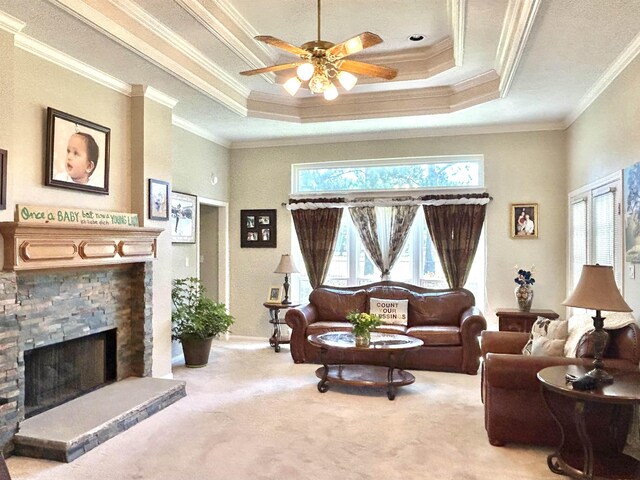 carpeted living room featuring a tray ceiling, a stone fireplace, ceiling fan, and ornamental molding