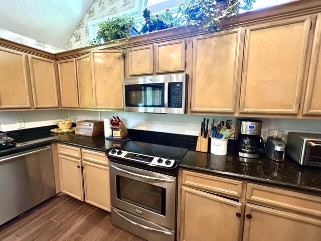 kitchen with appliances with stainless steel finishes, vaulted ceiling, dark wood-type flooring, light brown cabinets, and dark stone countertops