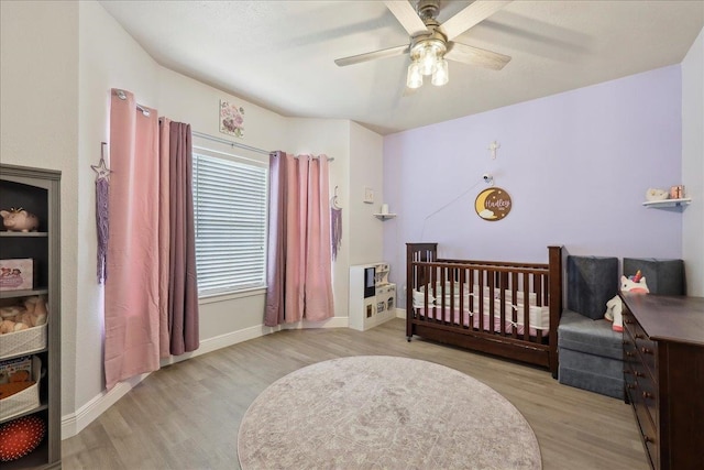 bedroom with light wood-type flooring, ceiling fan, a crib, and baseboards