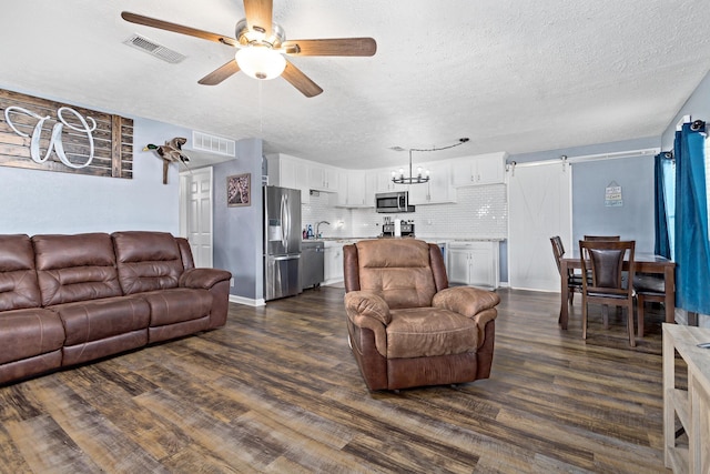 living room with a barn door, visible vents, dark wood-style floors, and ceiling fan with notable chandelier