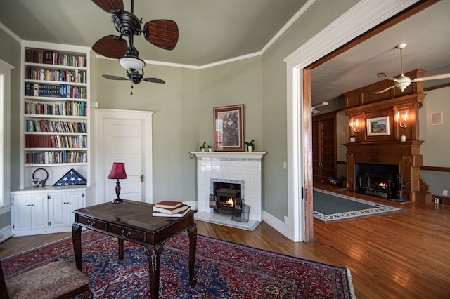 office with built in shelves, ceiling fan, wood-type flooring, and a tiled fireplace