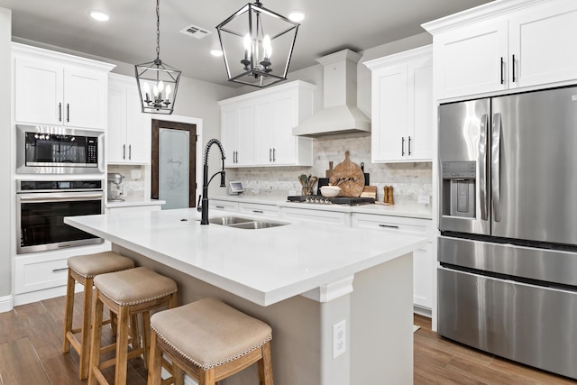 kitchen with white cabinets, appliances with stainless steel finishes, sink, a kitchen island with sink, and custom range hood