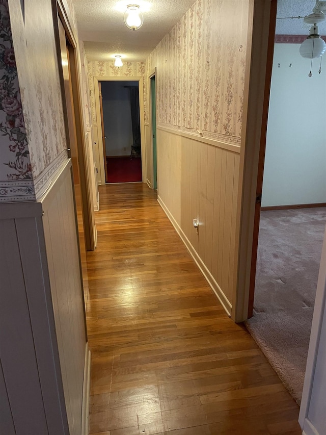 hallway featuring a wainscoted wall, a textured ceiling, wallpapered walls, and hardwood / wood-style flooring