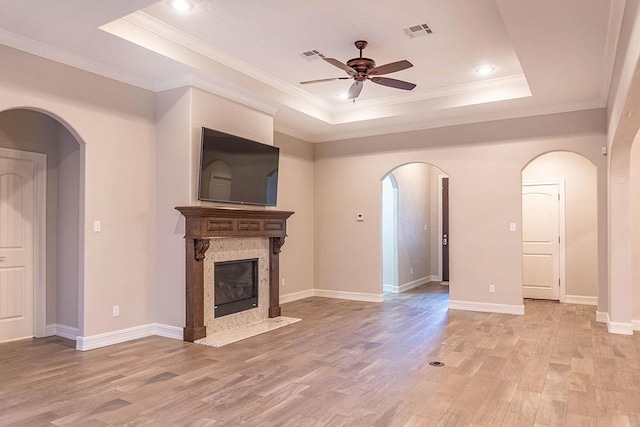 unfurnished living room with ceiling fan, a premium fireplace, ornamental molding, a tray ceiling, and light wood-type flooring