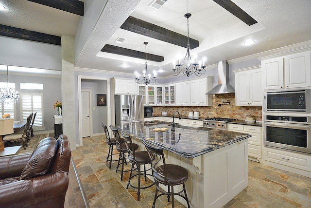 kitchen featuring stone tile flooring, appliances with stainless steel finishes, a raised ceiling, wall chimney range hood, and a notable chandelier