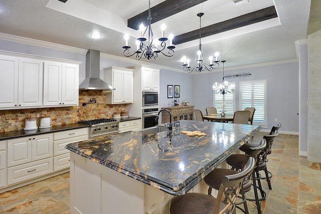 kitchen with dark countertops, a raised ceiling, an inviting chandelier, stainless steel appliances, and wall chimney exhaust hood