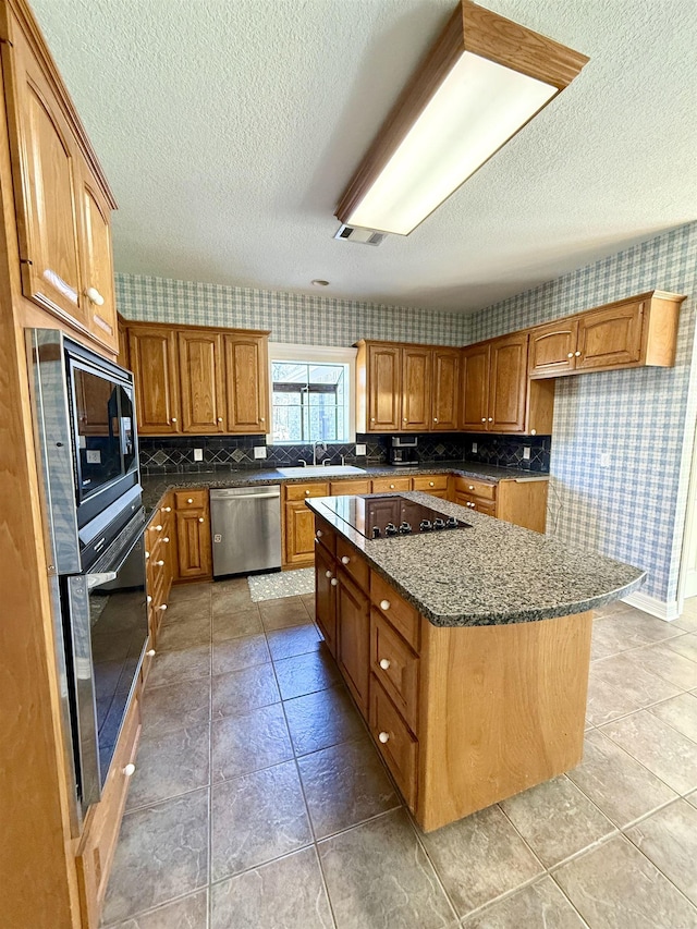 kitchen featuring a sink, dark stone countertops, stainless steel appliances, brown cabinetry, and wallpapered walls