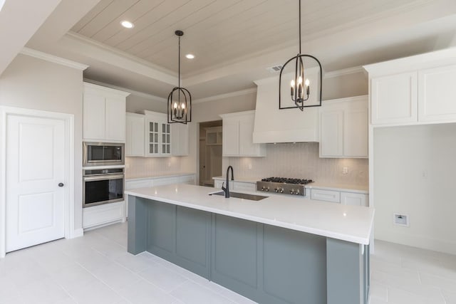 kitchen with pendant lighting, appliances with stainless steel finishes, a tray ceiling, white cabinets, and a center island with sink