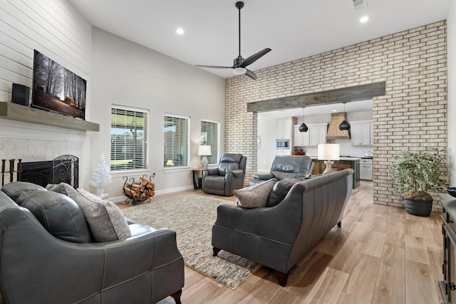 living room featuring ceiling fan, a high ceiling, brick wall, a tiled fireplace, and light wood-type flooring