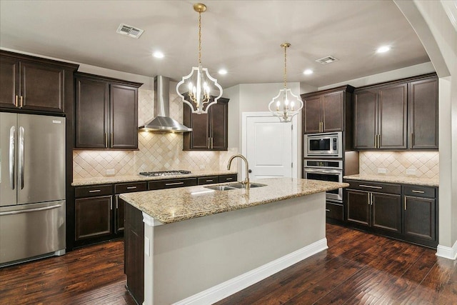 kitchen with light stone counters, stainless steel appliances, sink, wall chimney range hood, and a center island with sink