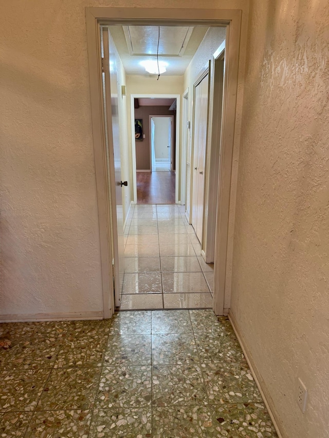 corridor with attic access, a textured wall, tile patterned floors, and baseboards