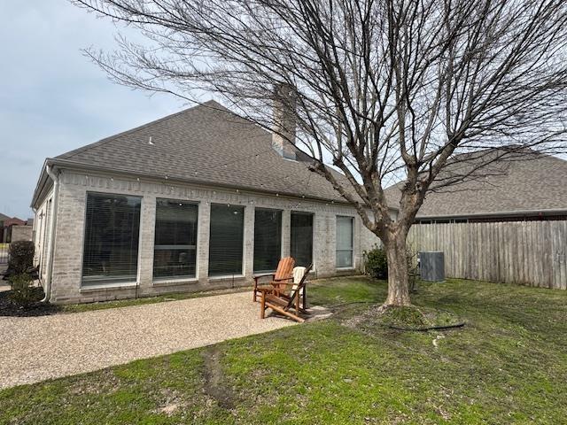 back of house featuring brick siding, fence, roof with shingles, a lawn, and a chimney