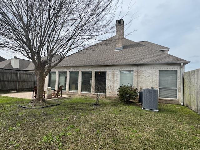 rear view of property featuring central air condition unit, brick siding, fence, a yard, and roof with shingles