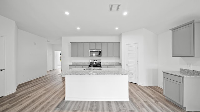 kitchen with gray cabinets, a kitchen island with sink, stainless steel appliances, and light stone counters