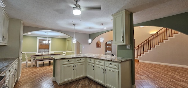 kitchen with light stone countertops, kitchen peninsula, pendant lighting, ceiling fan with notable chandelier, and ornamental molding