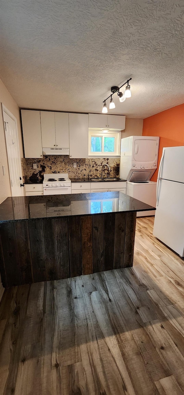 kitchen with dark countertops, white appliances, wood finished floors, under cabinet range hood, and stacked washer and clothes dryer