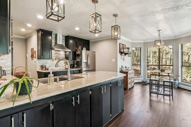kitchen featuring ornamental molding, stainless steel refrigerator with ice dispenser, dark cabinets, and wall chimney exhaust hood