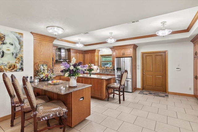 kitchen with wall chimney exhaust hood, stainless steel refrigerator with ice dispenser, a breakfast bar area, and a tray ceiling