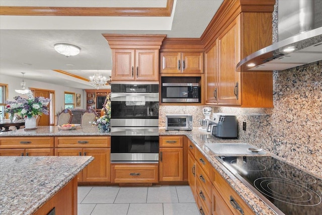 kitchen with appliances with stainless steel finishes, backsplash, wall chimney exhaust hood, a tray ceiling, and a chandelier