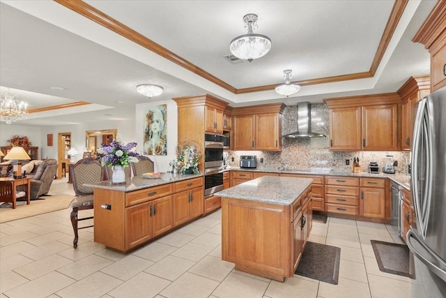 kitchen featuring a center island, wall chimney exhaust hood, an inviting chandelier, a tray ceiling, and appliances with stainless steel finishes