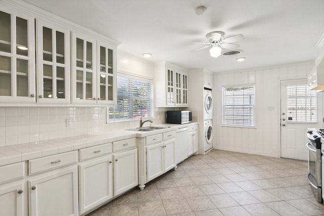 kitchen featuring tile countertops, stainless steel stove, white cabinetry, stacked washer / drying machine, and white dishwasher