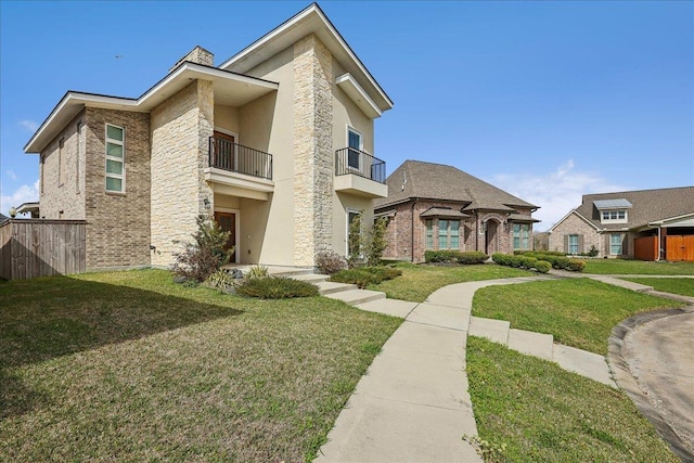 view of front facade with stucco siding, a balcony, fence, a front yard, and brick siding