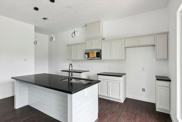 kitchen with a center island with sink, sink, hanging light fixtures, and dark wood-type flooring