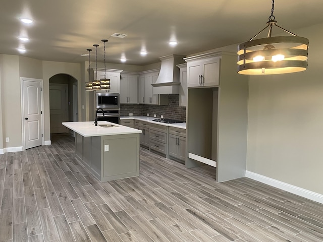 kitchen with custom exhaust hood, a center island with sink, decorative backsplash, appliances with stainless steel finishes, and decorative light fixtures